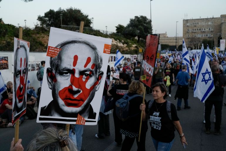 Антиправительственные протестующие маршируют в Израиле. Один из них держит плакат, на котором изображено лицо премьер-министра Биньямина Нетаньяху, обведенное красной рукой.