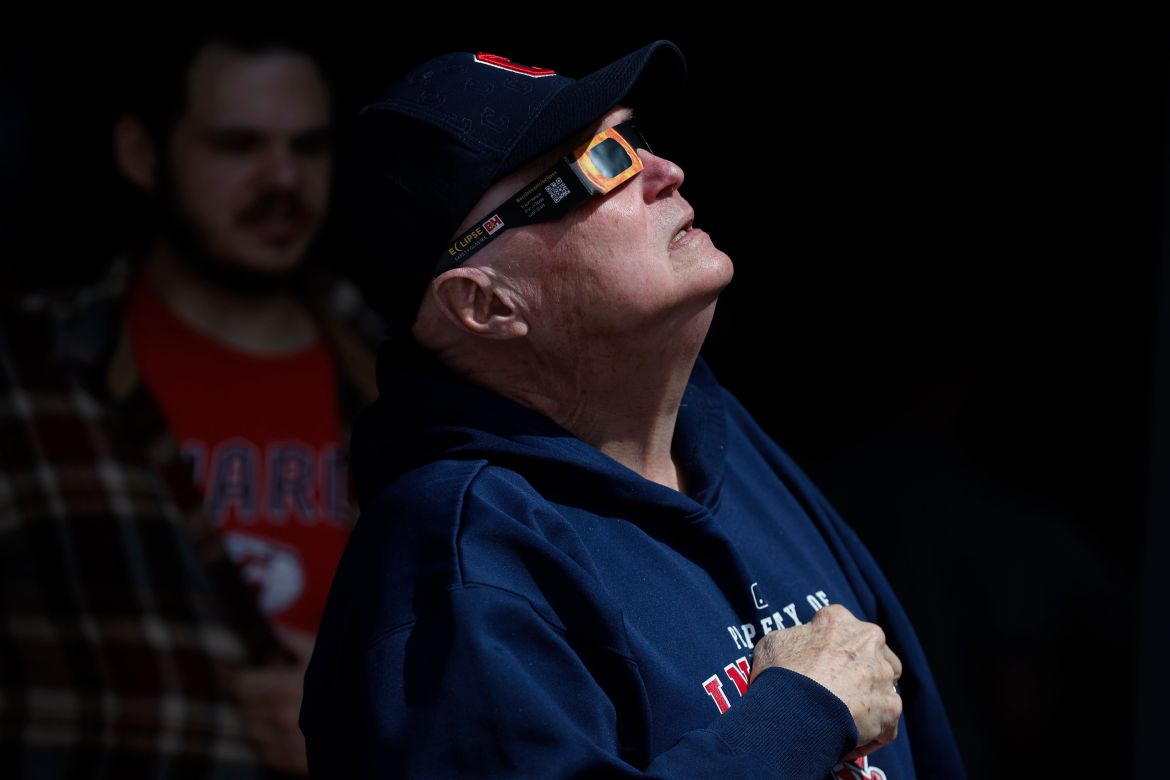 A man in a baseball cap and eclipse glasses looks up to the sky.
