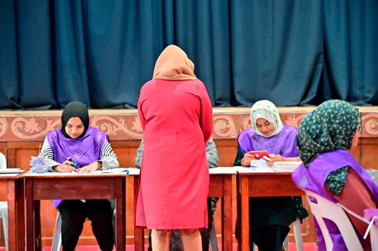 A voter casting her ballot in Madlives. She is wearing red and has her back to the camera. Elections officials in purple are facing her.