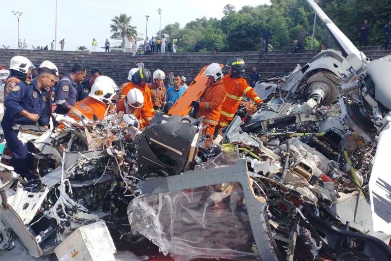 Rescuers looking through the wreckage of a navy helicopter that crashed in Malaysia. They are wearing orange high-viz suits. People in naval uniform are also at the crash site. There are steps behind, and people standing on the top.