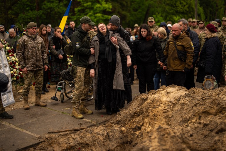 Two men support an older woman at the funeral for Ukrainian army paramedic Nazarii Lavrovskyi. Peopel are gathered behind. some in uniform and others in black. One has a Ukraine flag. There is a pile of earth to the right of the picture. 