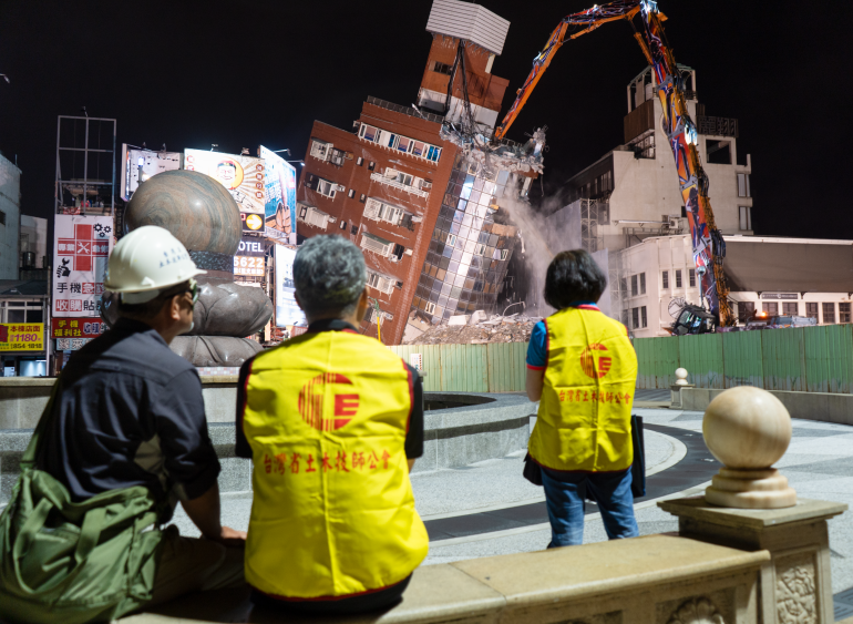 A collapsed building in Hualien being taken down by an excavator. People in yellow high-vis jackets are watching from a distance.