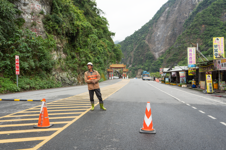 A worker in front of the entrance gate to the Taroko Gorge National Park. There are steep cliffs. Bollards have been placed to cordon off the road.