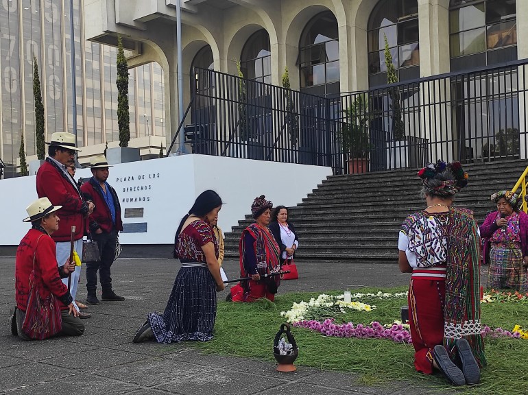 Women kneel for a ceremony outside of a courtbuilding in Guatemala City. A ring of flowers sits in front of them.