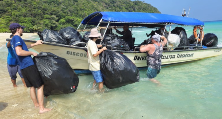 A group of men in shorts and T-shirts loading large lakc rubbish bags into a small boat near the beach. The sea is clear. 