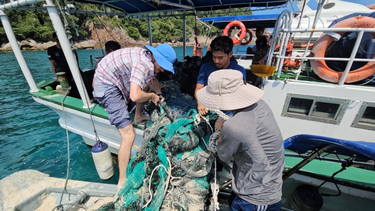 Группа людей переносит огромную путаницу выброшенных рыболовных сетей на небольшую лодку. Сети зеленые и серые, узловатые. 