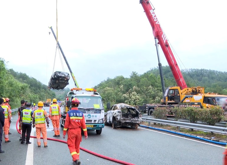 Rescuers at the scene of the collapse. A car is being lifted on a crane above the carriageway. Other recovered cars are already on the road. People in orange unifomrs and hard hats are nearby 