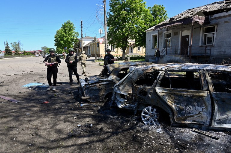 Ukrainian police inspect cars destroyed by a Russian bomb in Zolochiv. The cars are parked alongside the road but burned out. The roof of a building next to them has been blown off and the windows blown out