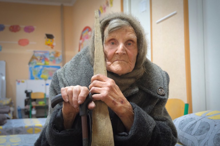 Lidya Stepanovna. She is sitting on a bed with a shawl around her head and leaning on a cane.