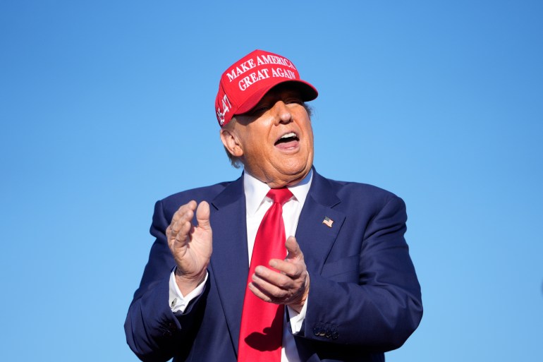 Donald Trump claps against a wide   bluish  sky, wearing a reddish  MAGA ballcap.