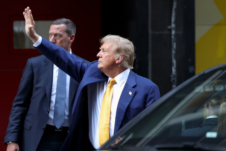Donald Trump, dressed in a blue suit and yellow tie, waves as he enters a vehicle.