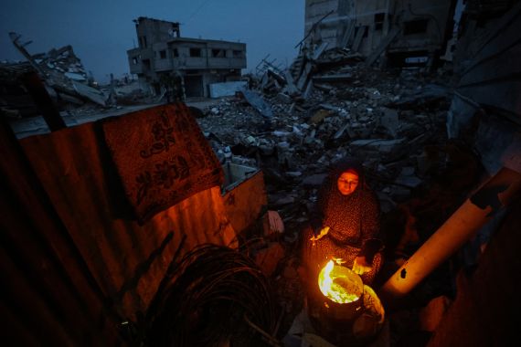 A displaced Palestinian woman warms herself beside a fire amid the ruins of destroyed buildings in the Bureij refugee camp, in the central Gaza Strip