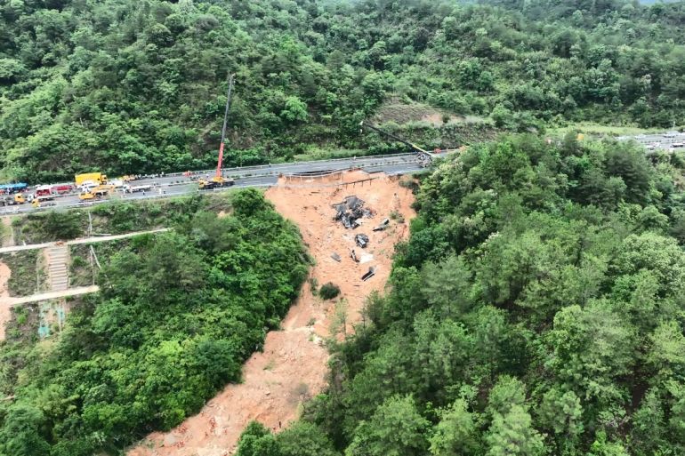 An aerial view of the disaster site. One side of the road has collapsed down a hill. The path left by the collapse is shown by brown earth cutting through the trees