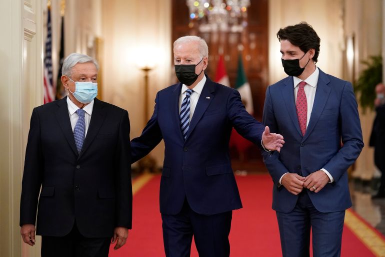Three leaders walking on a red carpet in what looks to be the White House - with Biden showing them around, as he has his arms wide open as if he is explaining the surroundings. All three are wearing facemasks