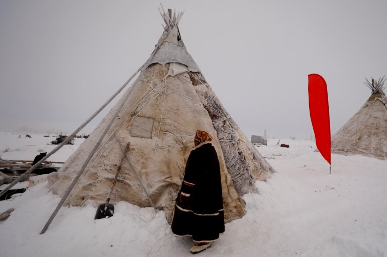 An image of a Nenet woman standing in front of a tent in a snowy area with another tent on the side of the photo in the Obdorskiy Ostrog , center Salekhard town of Yamalo-Nenets autonomous district, Russia