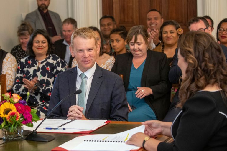 Chris Hipkins is sworn in as Prime Minister. He is sitting at a table with Governor General Cindy Kiro who is on the right of frame. They have papers on the table in front of them and there is a flower arrangement in the centre. There are people watching, most of them are women. They look happy