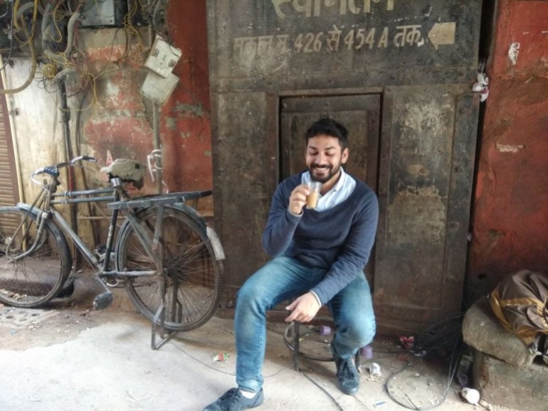 smiling young man drinking a cup of chai, seated on a stool in what looks like an alleyway