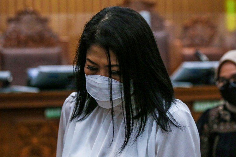 Putri Candrawathi in court. She is wearing a white shirt and white face mask. Her head is bowed so her hair slightly covers her face.