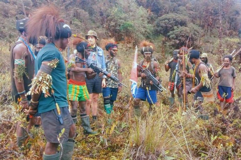 A man said to be Phillip Mehrtens in a line with armed West Papua rebels, The rebels are in shorts, t-shirts and wellington boots. Some are wearing traditional hats with plumes and have leaves and grasses tied to their biceps. There is a West Papua independence flag behind them.