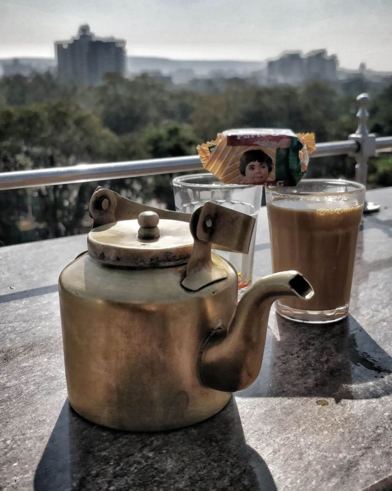 A teapot with two cups, one filled with chai and the other empty, with a packet of biscuits balanced on them