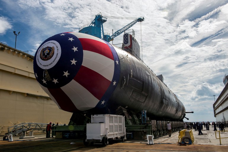 A Virginia-class submarine on the dry dock at a shipbuilding yard. Its nose is decorated in the colours of the United States. There is a crane behind. It dwarfs the people that are milling about around it.