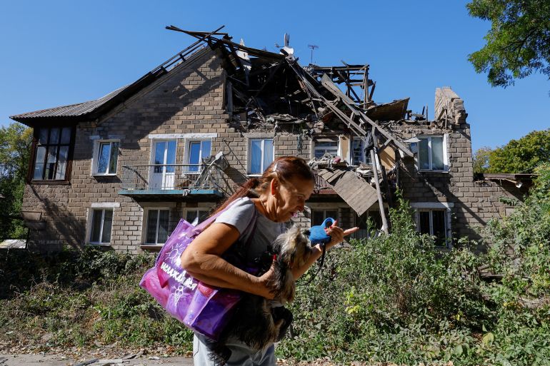 A woman walks past a shelled house in a Russian-occupied part of Donetsk. She is holding a white dog.