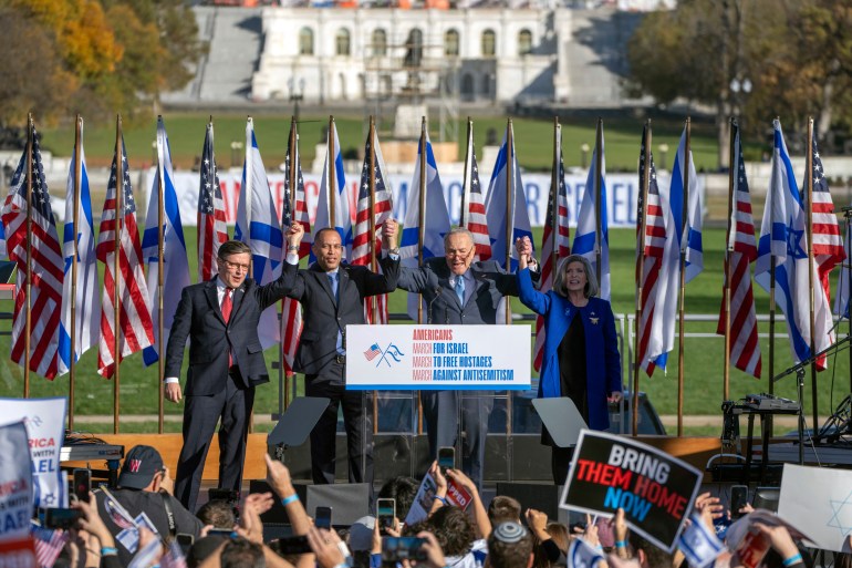 Speaker of the House Mike Johnson of La., left, House Minority Leader Hakeem Jeffries of N.Y., Senate Majority Leader Chuck Schumer of N.Y., and Sen. Joni Ernst, R-Iowa, right, join hands at the March for Israel. There are banners behind them and people in front