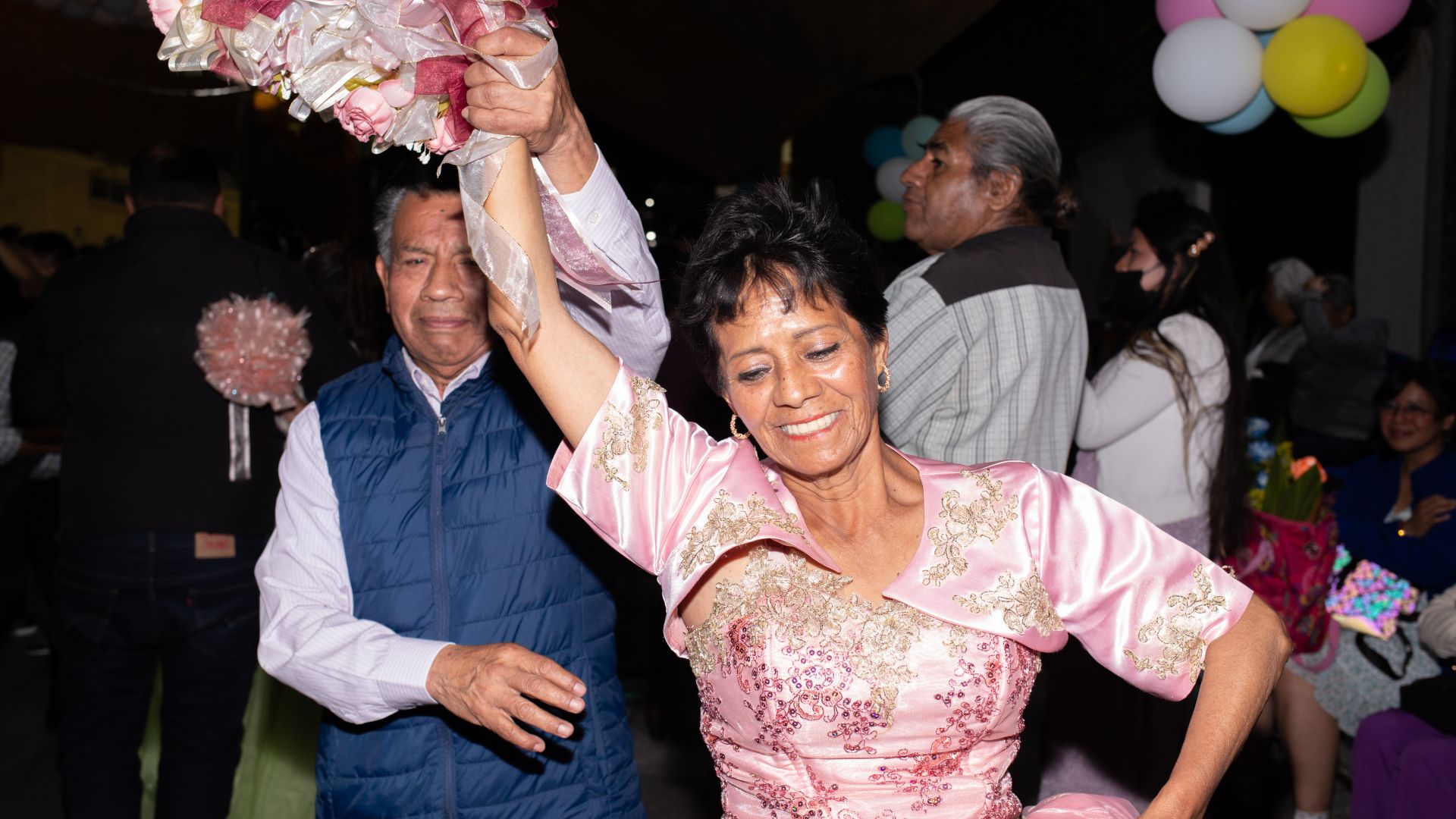 A woman in a pink dress is twirled by her dance partner, one arm lifted into the air as she perches the other on her hip.